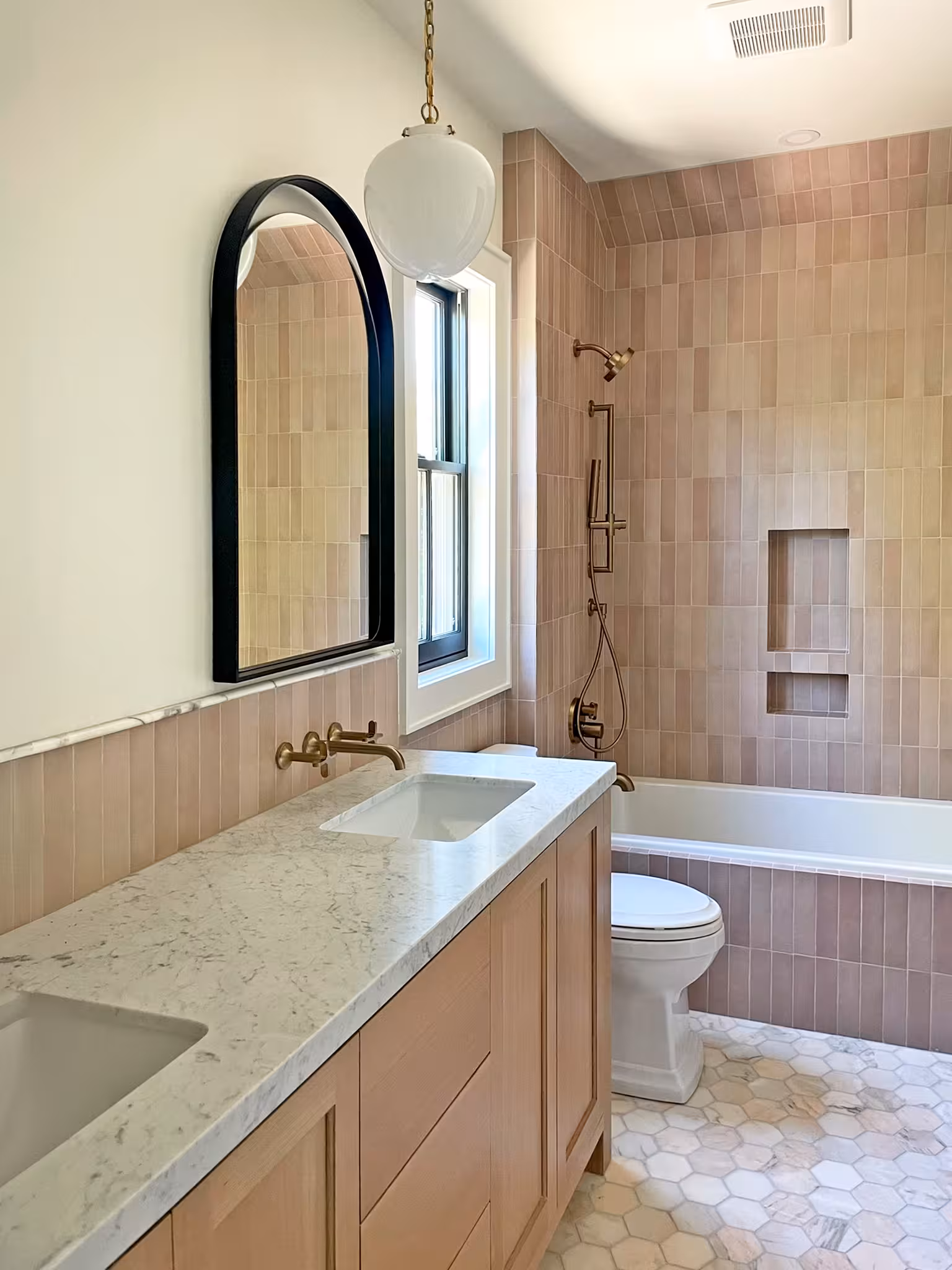 Holladay home bathroom with a light wood vanity, pink vertical tile and an arched black mirror