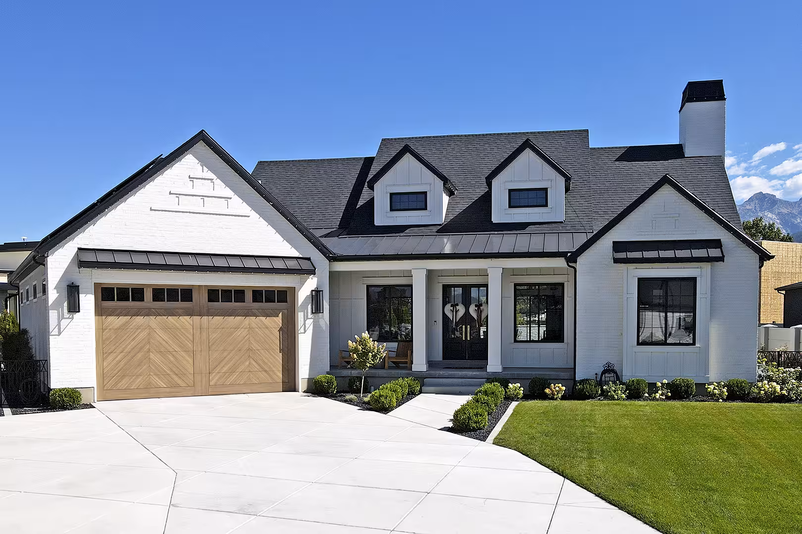 Front of the Meadow home with white brick, wood garage doors and black metal porch roofs